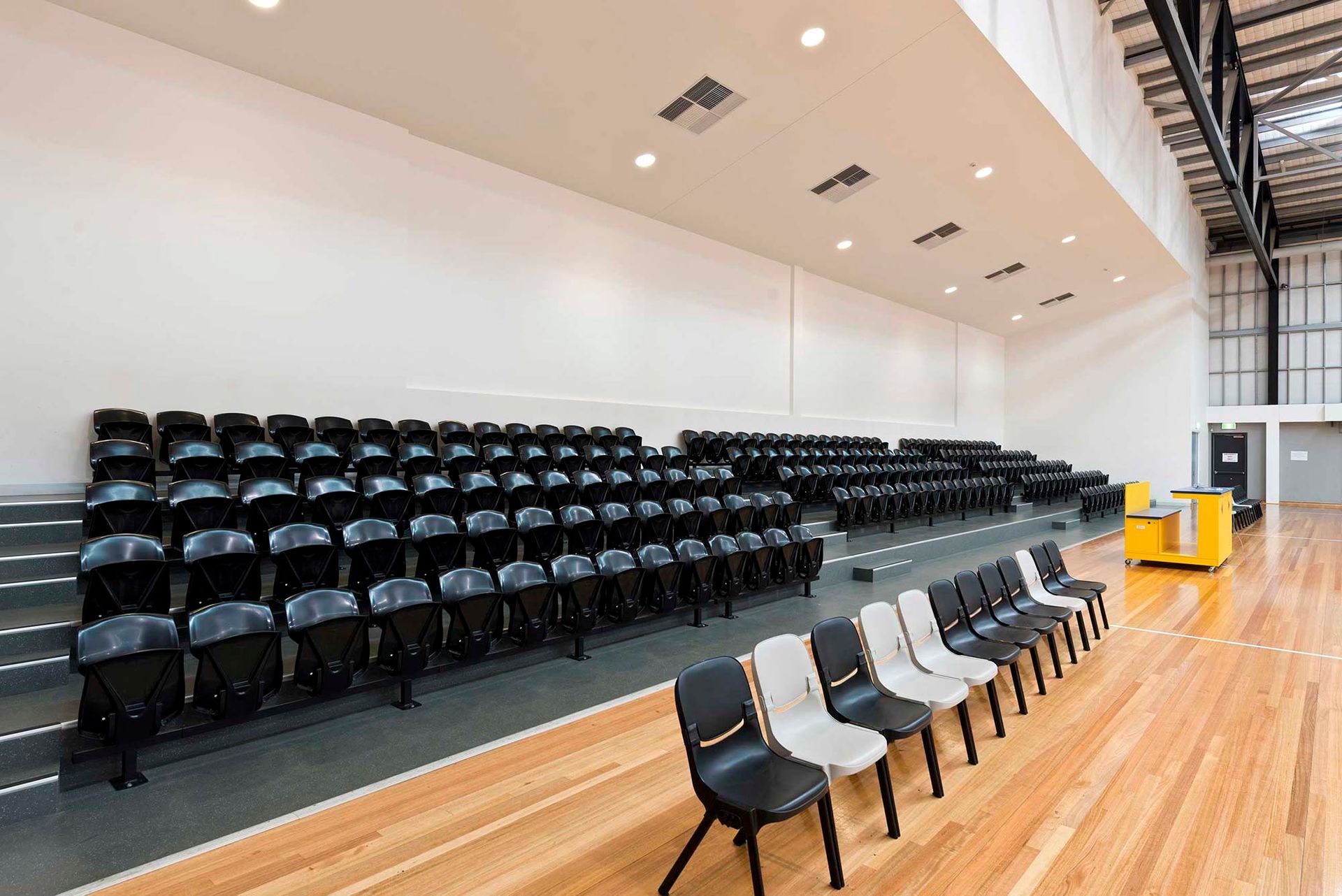 A large auditorium with rows of black chairs on a wooden floor.