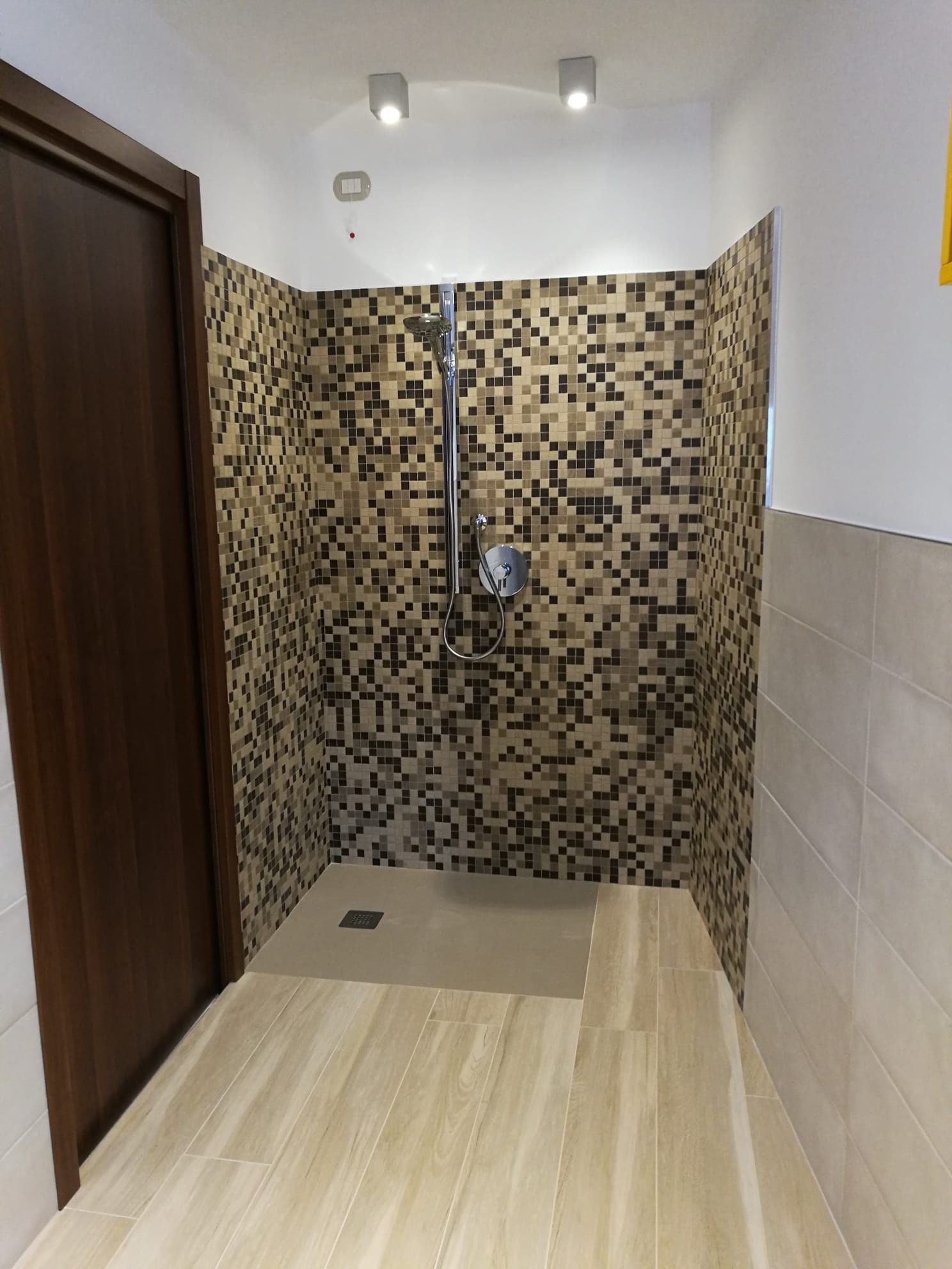 Piccola zona doccia piastrellata con pavimento beige e piastrelle a mosaico marroni alle pareti, vista dalla porta.