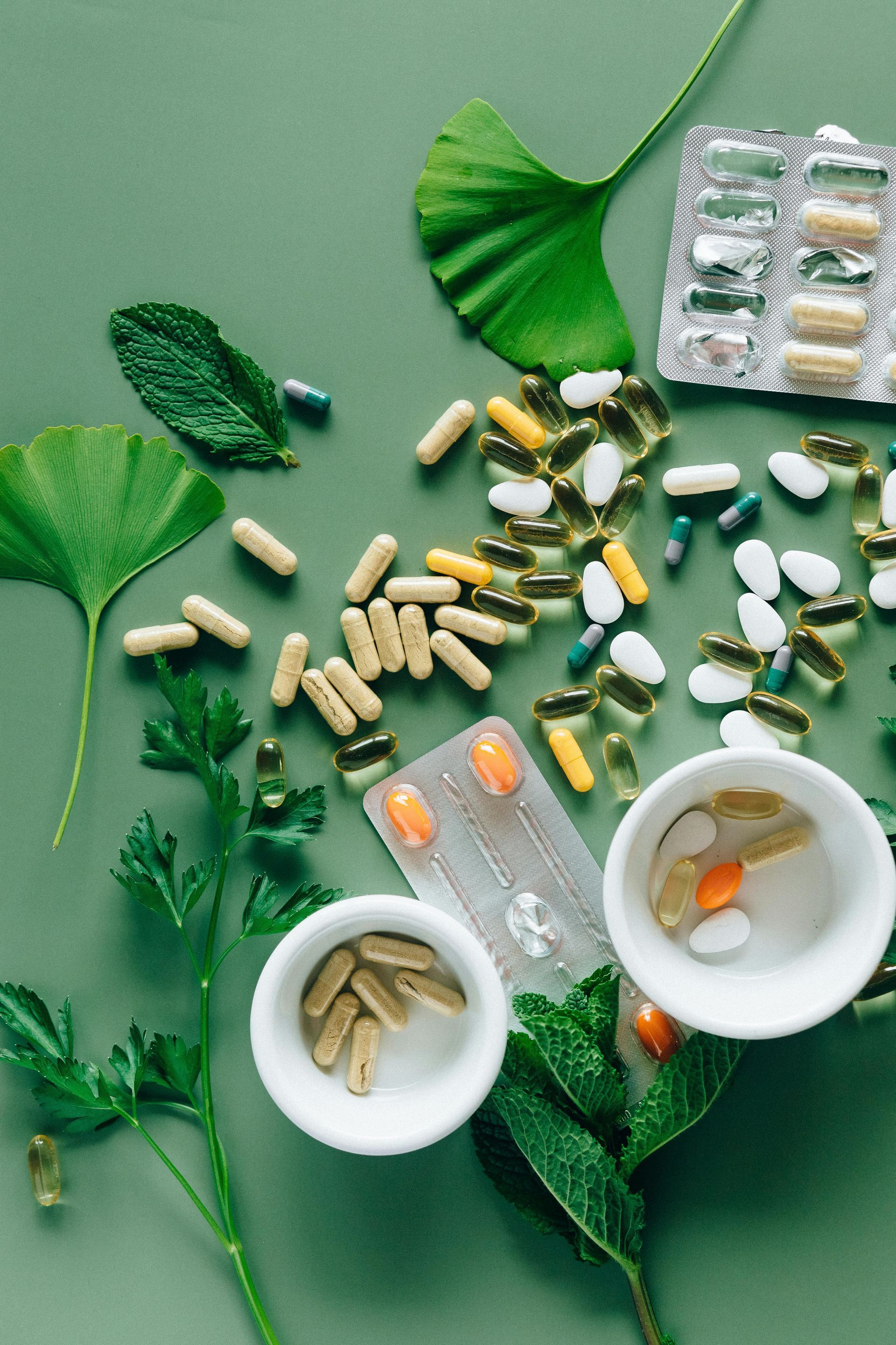 Assortment of pills and capsules scattered on a green surface, alongside green leaves and small white bowls.