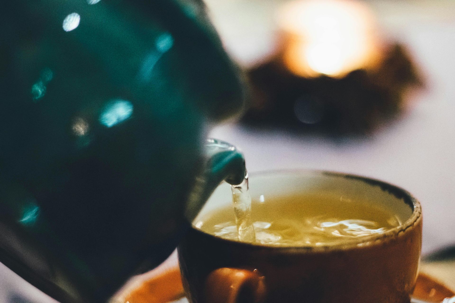 Green teapot pouring tea into an orange mug, with a blurred lit candle in the background.