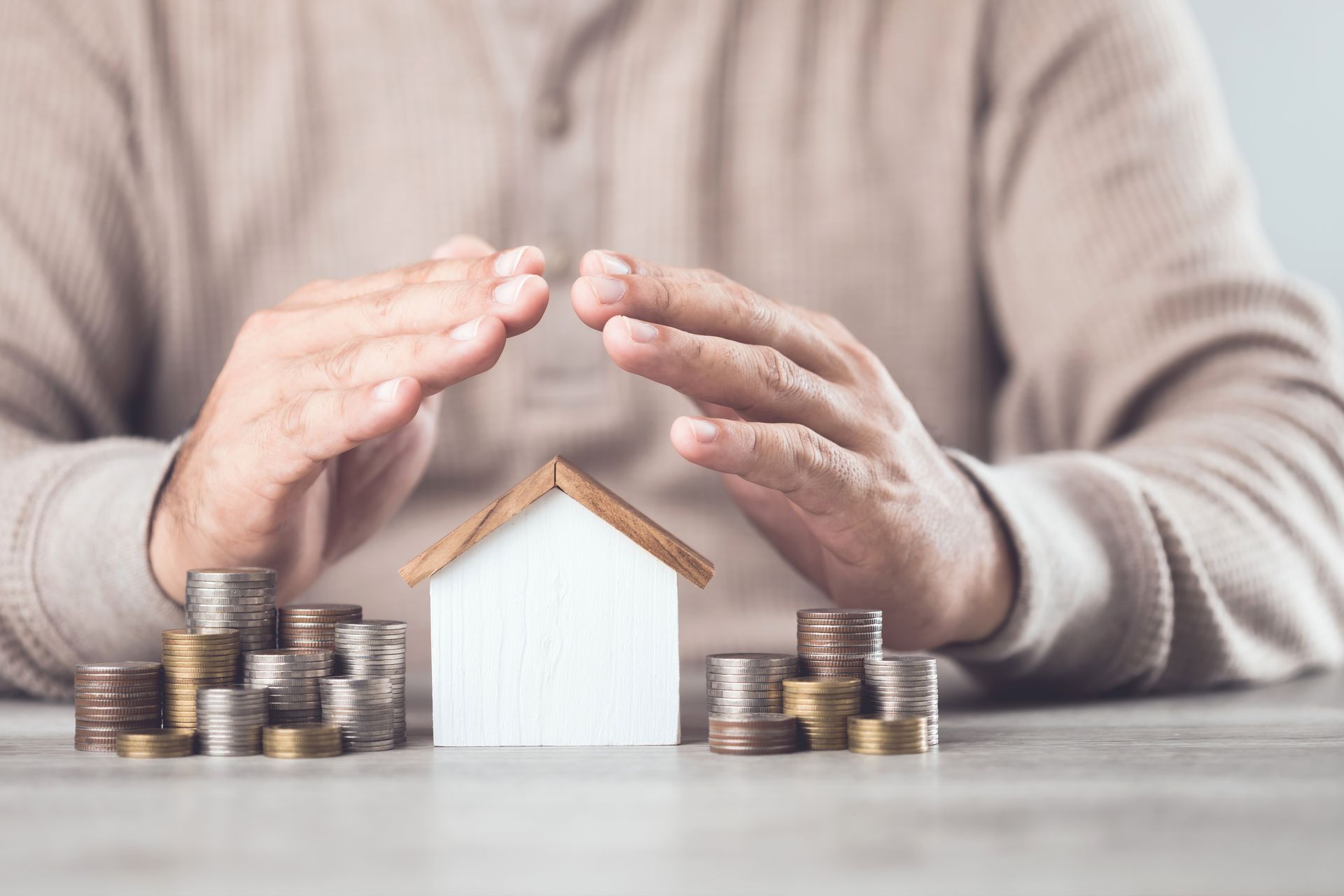 Hands protecting a small house and stacks of coins, suggesting financial security.