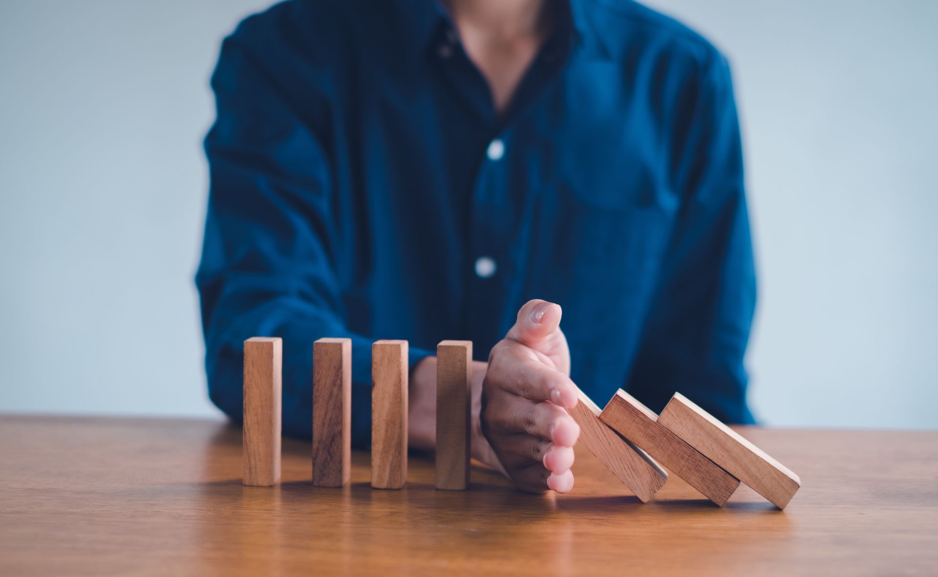 Person stopping dominoes from falling, blue shirt, wooden table.