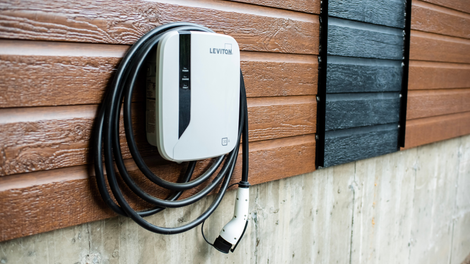 White EV charger mounted on a brown and black siding wall, with a black charging cable and plug.