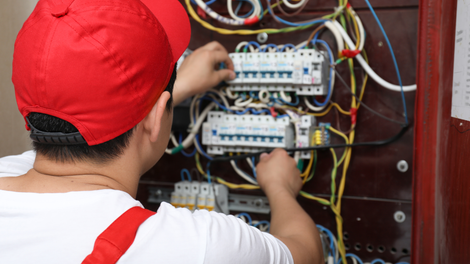 Electrician in red cap and white shirt working on electrical panel with wires.