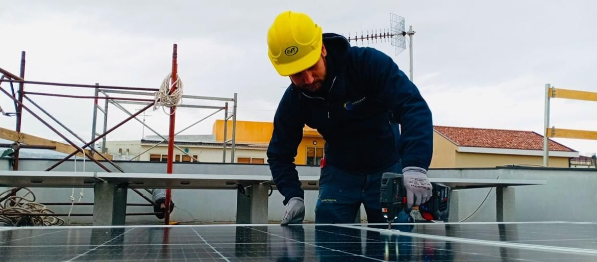 Due operai installano pannelli solari su un tetto di metallo rosso sotto un cielo azzurro.