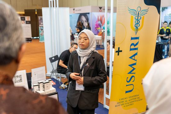 Woman in hijab speaks at USMARI booth, likely a medical and health fair, with a yellow banner.