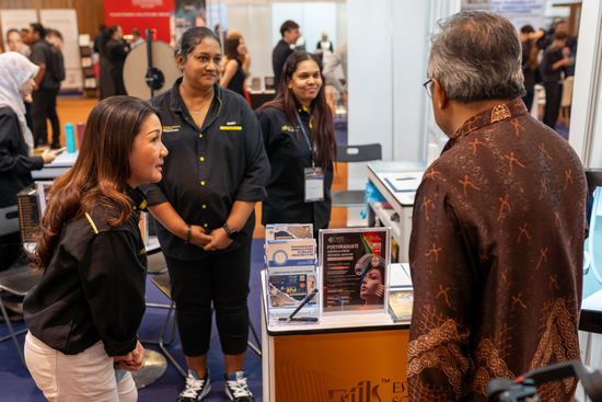 People at a trade show booth, talking. One person in patterned shirt faces booth. Two staff in black shirts.
