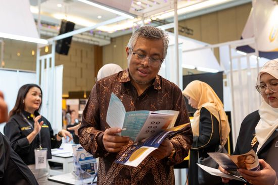 Man in patterned shirt looking at brochures at an event. Other people are in the background.