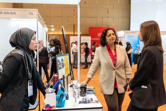 Three women converse at a booth during an event. One in a hijab, one in a blazer, and another in a suit.