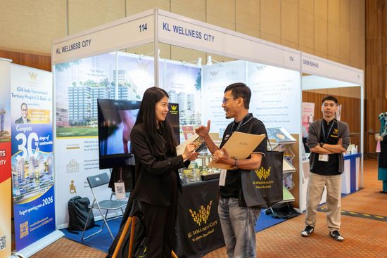 Woman and man shaking hands at a booth in an event hall, talking, with a third person looking on.