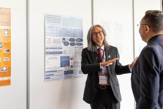 Two men at a conference presentation. One points to a poster, explaining content to the other, who listens attentively.