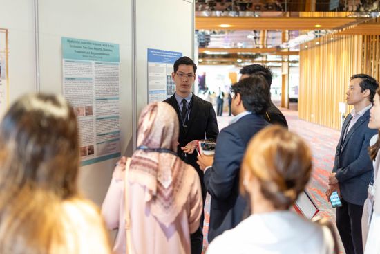 People gather at a poster presentation. A man in a suit gestures as he speaks, explaining a poster to a small audience.