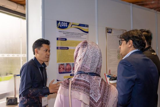 Man presenting a poster at a conference. Two people listen, one wearing a hijab. Setting: conference hall.