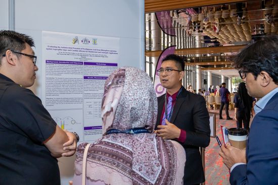 People at a conference, looking at a poster presentation. A man in a suit speaks, gesturing. Others listen, holding a coffee cup.