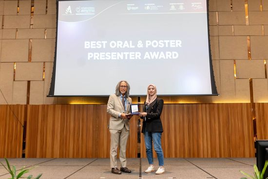 Man in suit presents award to woman on stage; banner reads 