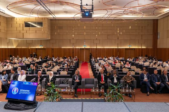 Conference hall with panelists on stage, audience seated. Red carpet, screen, and projector visible.