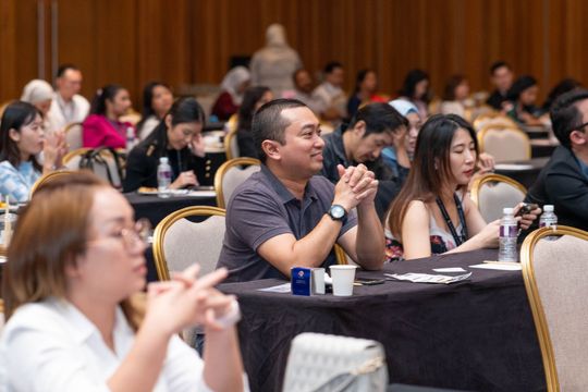 Attendees at a conference in a large room, some with hands clasped, looking forward.