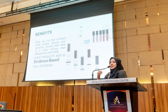 Woman in hijab presenting at a podium; a projector screen displays charts and text with the word 