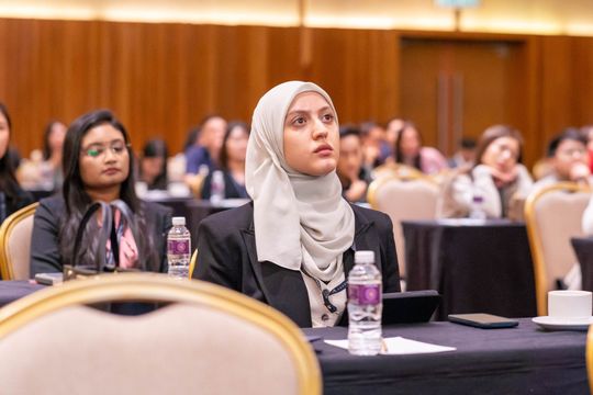 Woman in hijab at a conference, looking up intently, with other attendees in the background.
