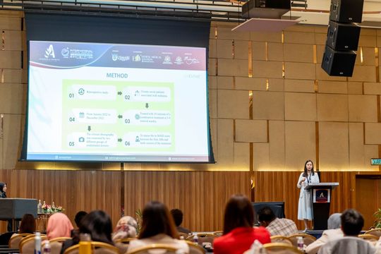 Woman presenting at a conference, large screen showing a diagram. Audience seated in front.