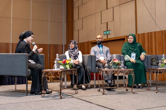 Panel discussion: Four people seated on a stage, speaking. Woman at left holds a microphone.