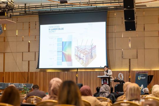 A presenter standing at a podium giving a presentation on a screen with diagrams. Attendees are seated in the audience.