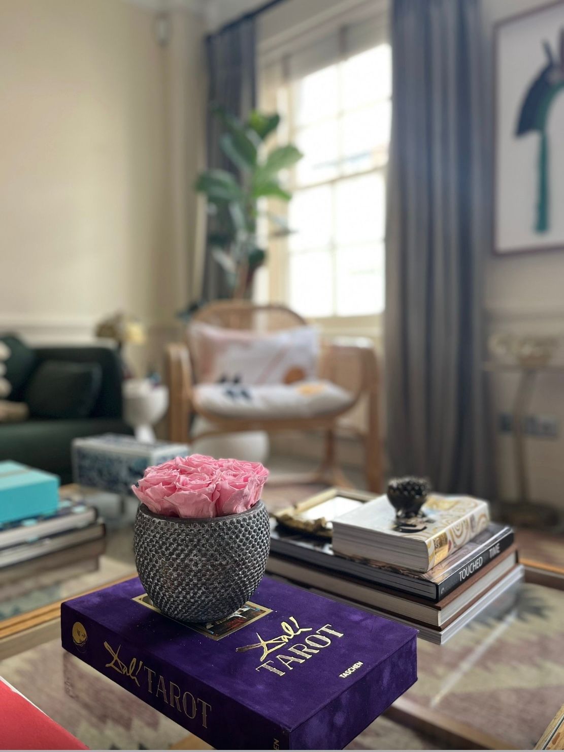 Coffee table with books, a vase of pink preserved flowers, and a living room in the background.
