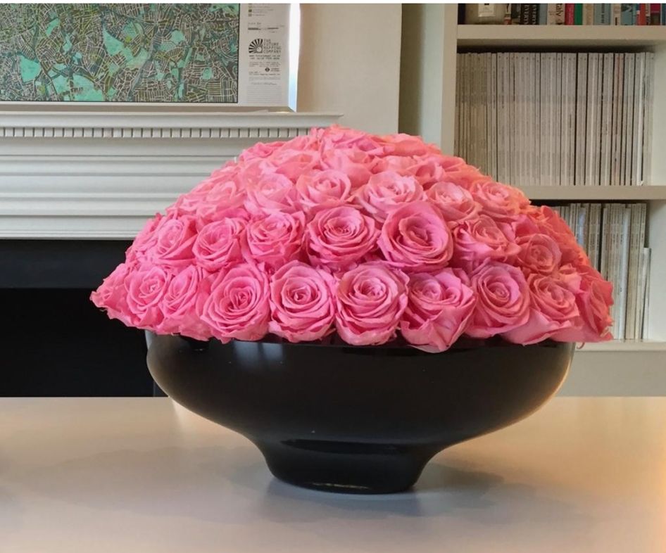 Pink roses in a black bowl on a table; background: white mantel, bookshelves.