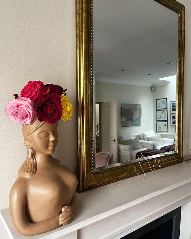 Woman-shaped vase with roses, golden mirror reflecting living room, on mantelpiece.