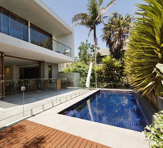 Modern house with pool; wooden deck, glass railings, and tropical foliage.