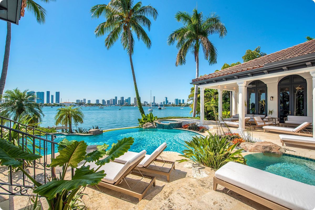 Luxury waterfront home with pool, lounge chairs, and palm trees; city skyline in the distance.