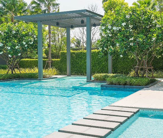 A swimming pool with stepping stones, a pergola, and trees on a sunny day.