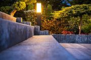 Lit stone walkway beside a glowing garden lamp and low wall at dusk
