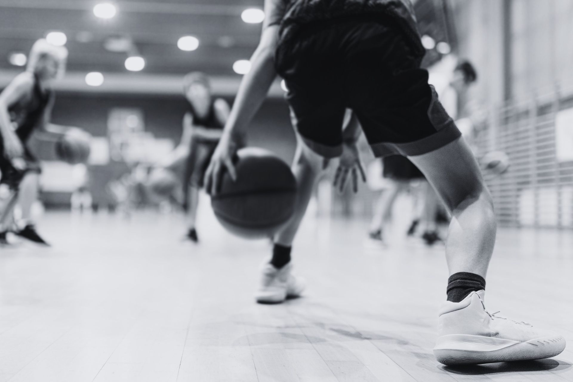 Basketball player dribbling the ball on a wooden court, other players in the background. Black and white.