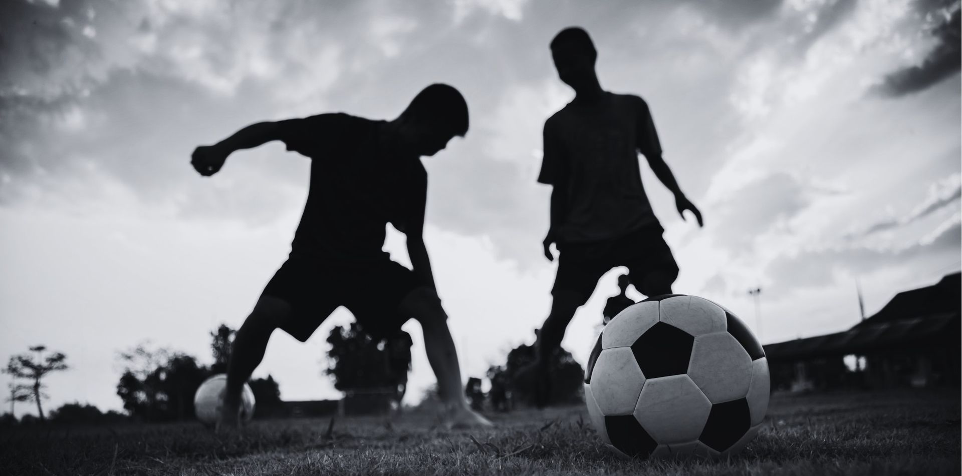 Two silhouetted figures playing soccer on grass under a cloudy sky.