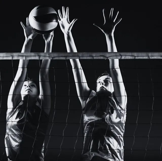 Two volleyball players jumping to block a ball over a net, in black and white.