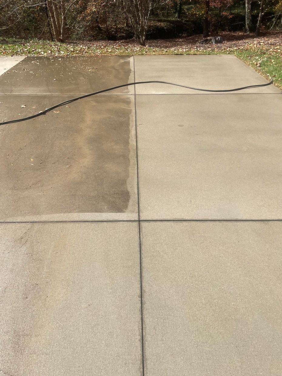 A concrete driveway with a stark contrast between a dirty, dark gray section being cleaned and a clean, light gray area.