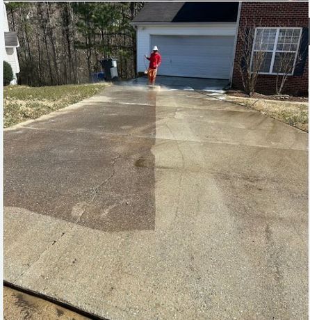 A person in high-visibility gear power washes a concrete driveway, showing a clear contrast between dirty and clean areas.