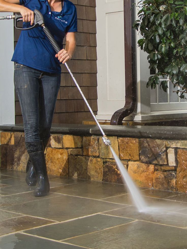 A person in a blue polo and dark pants uses a pressure washer to clean a stone patio next to a house exterior.