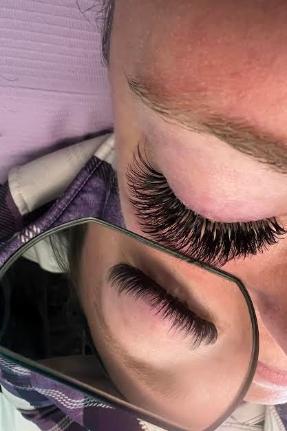 Woman receiving facial treatment in a spa-like setting; esthetician's hands near her face. Bright overhead lights.