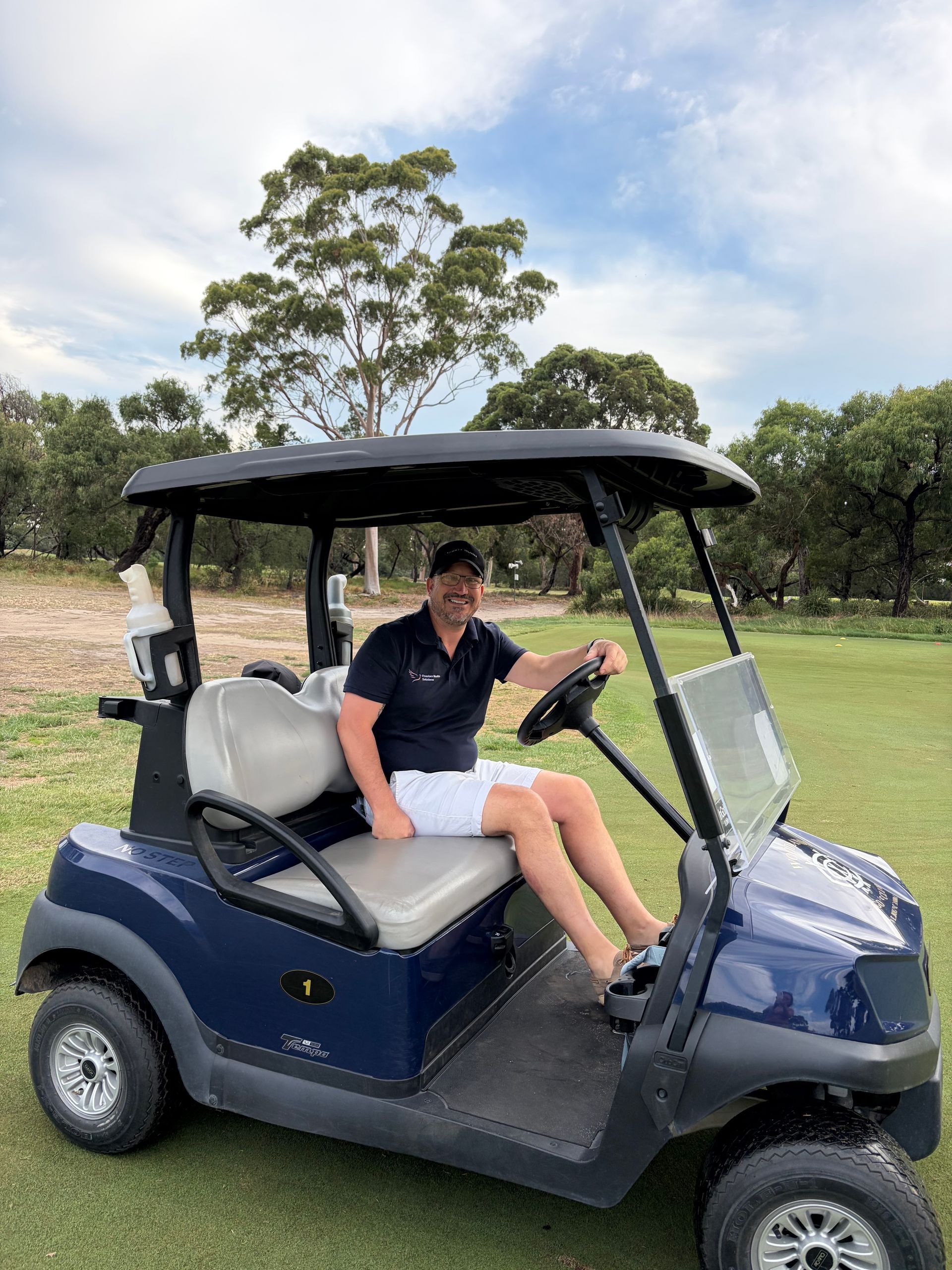 A person in a dark shirt and white shorts sits in a navy-blue golf cart on a green course with trees in the background. — Freedom Wealth Solutions in Mornington, VIC