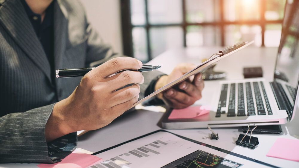Person in Suit Writing on Clipboard at A Desk with A Laptop — Freedom Wealth Solutions in Mornington, VIC