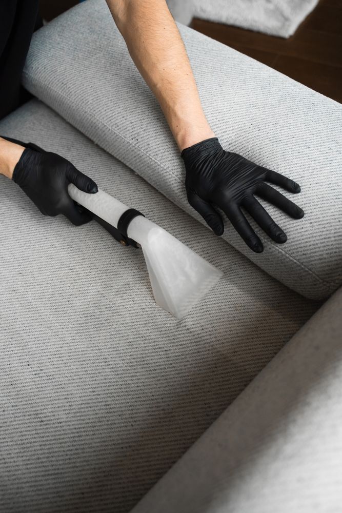 A person wearing black gloves is cleaning a couch with a vacuum cleaner.