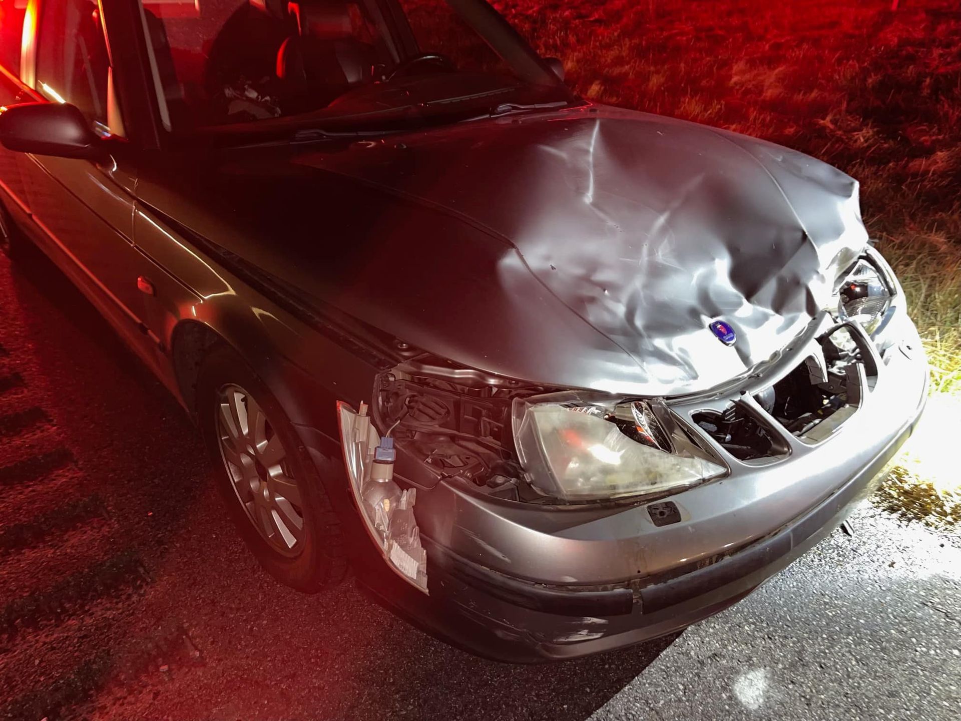 A silver car with a damaged hood is parked on the side of the road