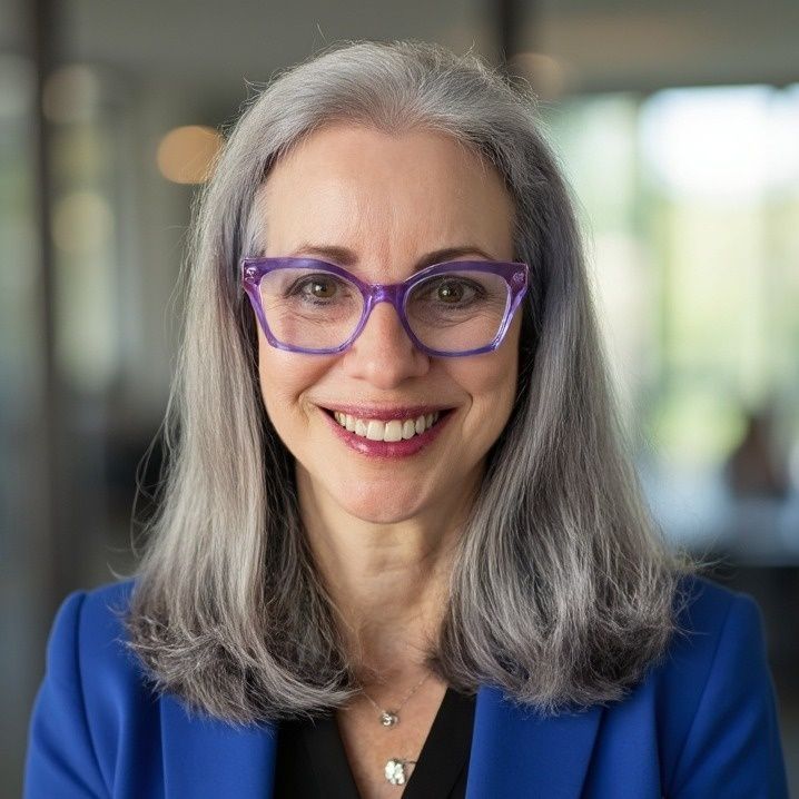 Woman with gray hair and purple glasses smiling, wearing a blue blazer.