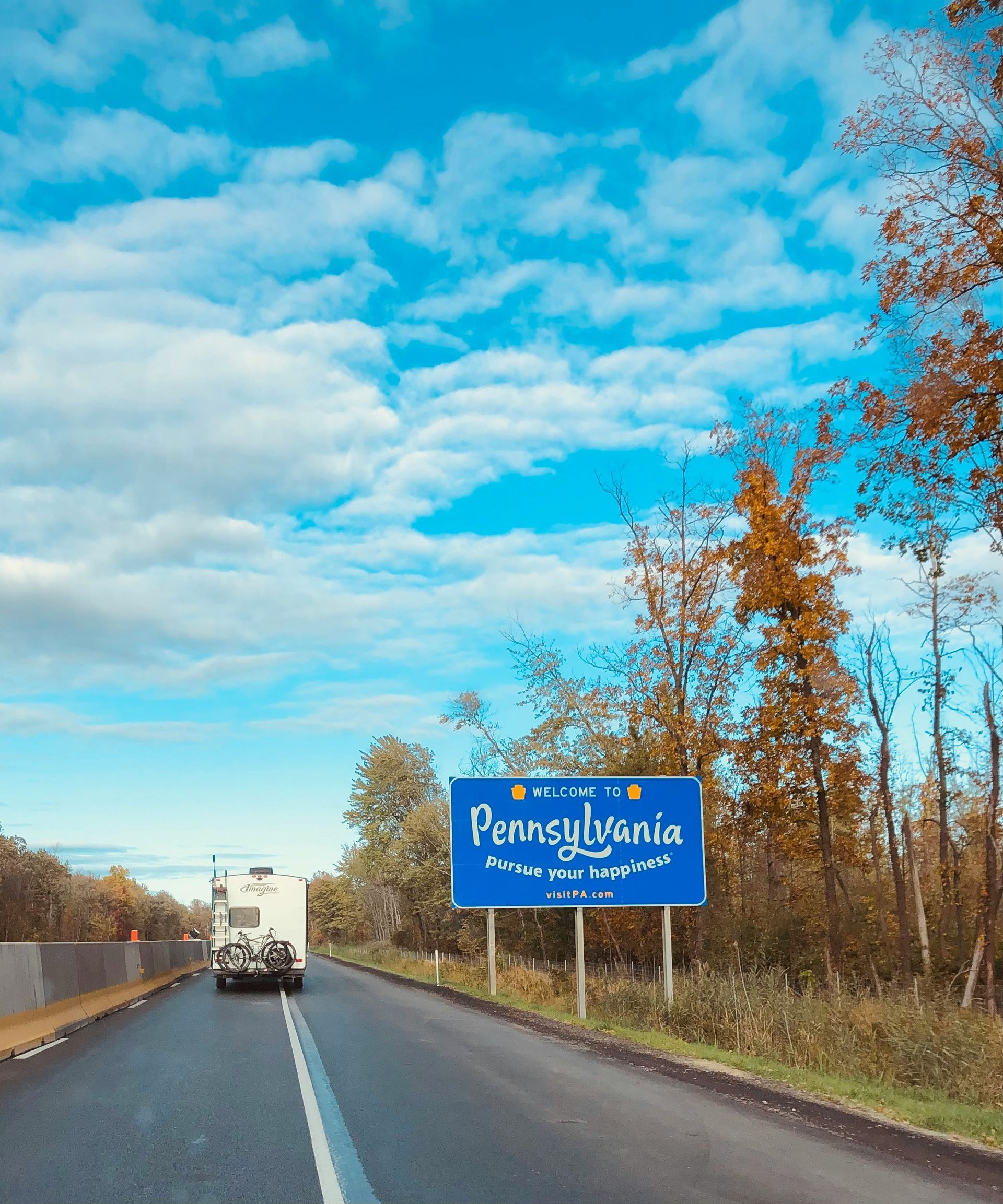 Pennsylvania welcome sign along a road with a RV, blue sky and autumn trees.