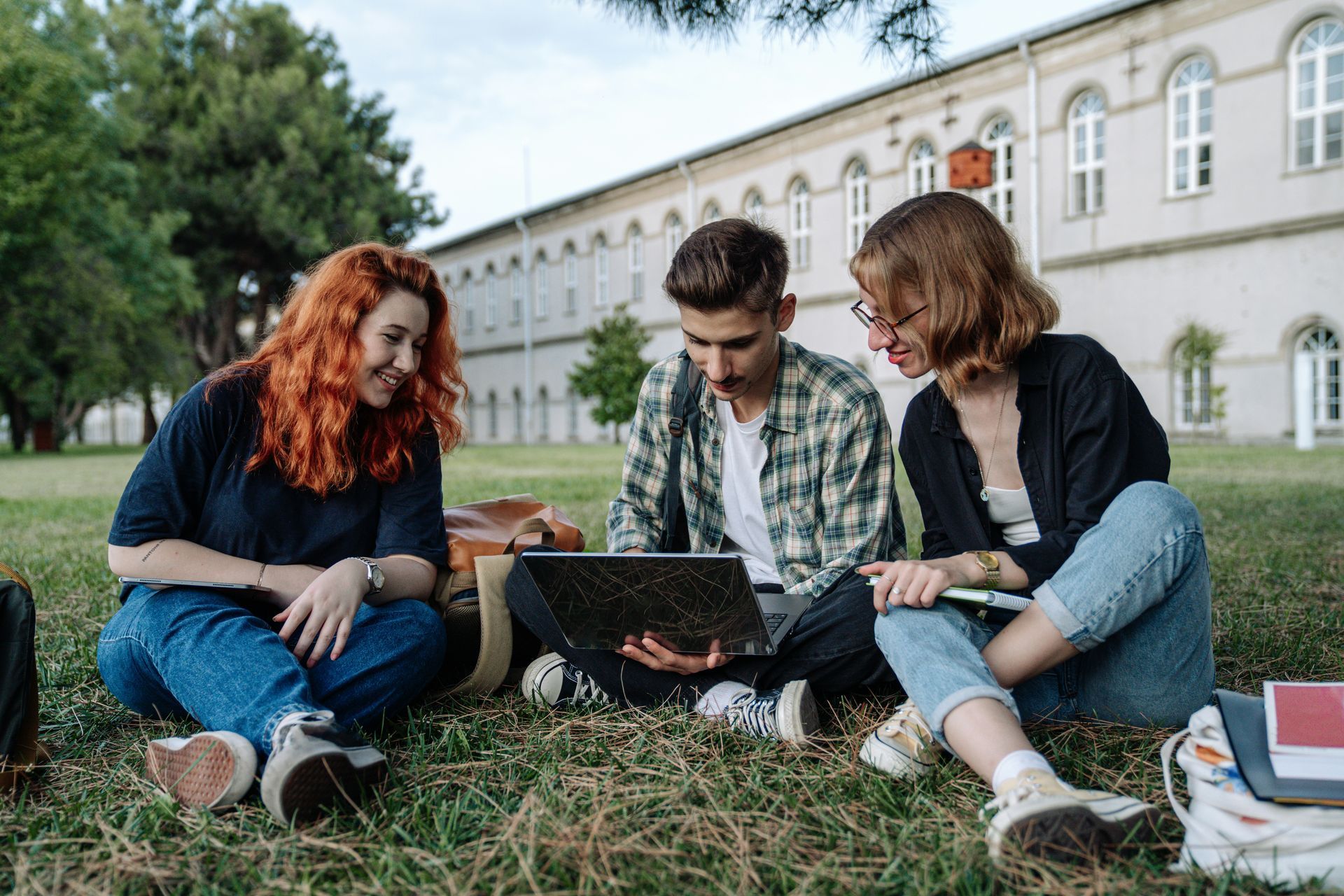 Three people sit on grass looking at a laptop. A large building is in the background.