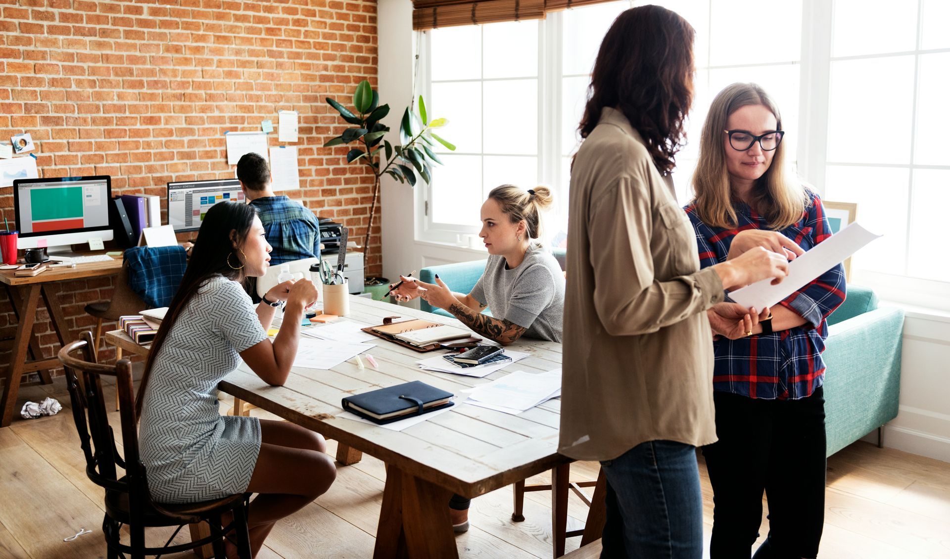 Office meeting with several people around a table and working at desks in a brick-walled room.