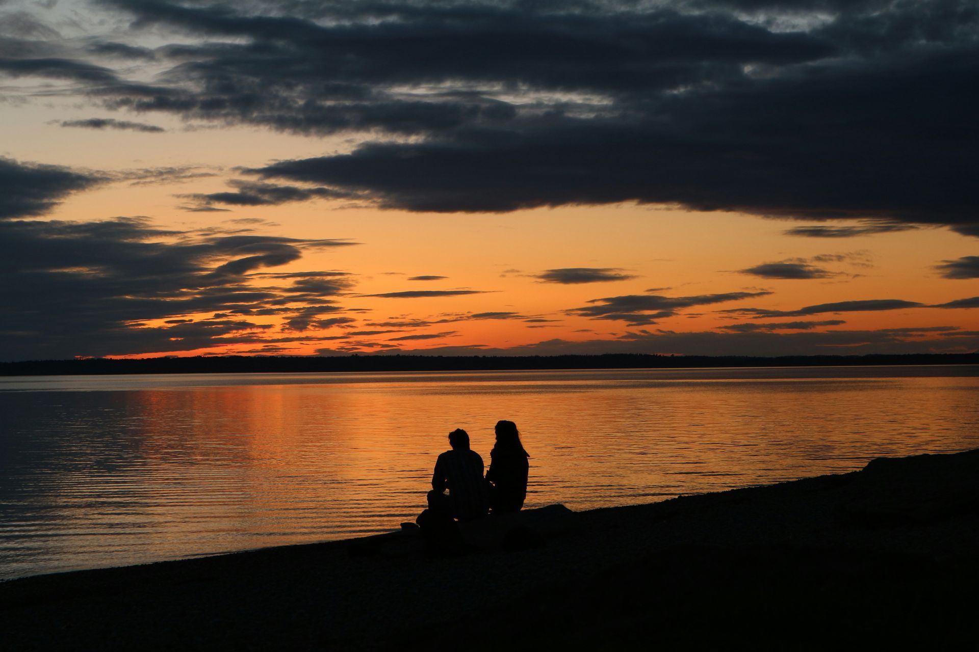 Silhouetted figures watching sunset over a calm body of water; orange and gray sky.
