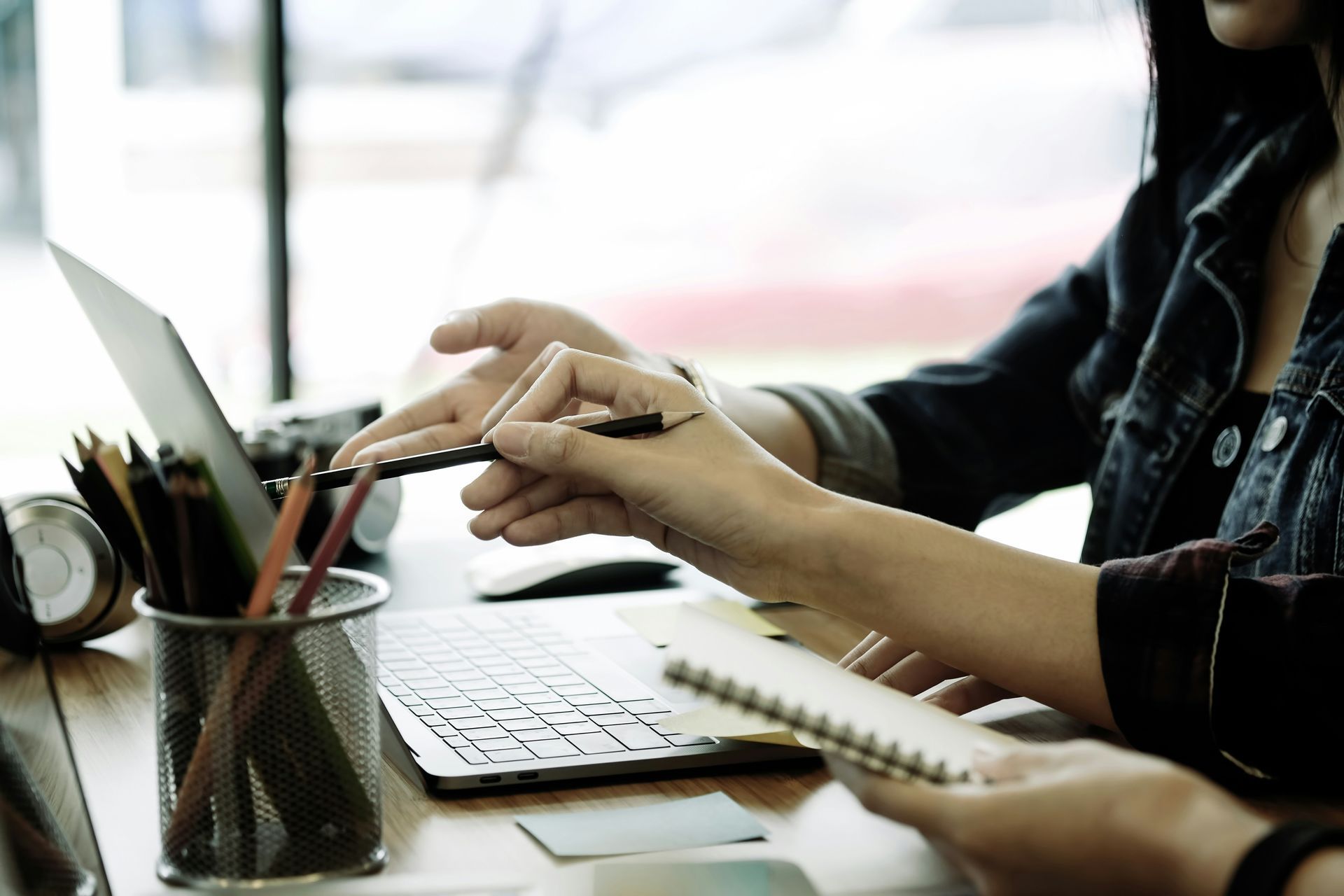Two people collaborating at a desk, looking at a laptop and notepad, using a pen.